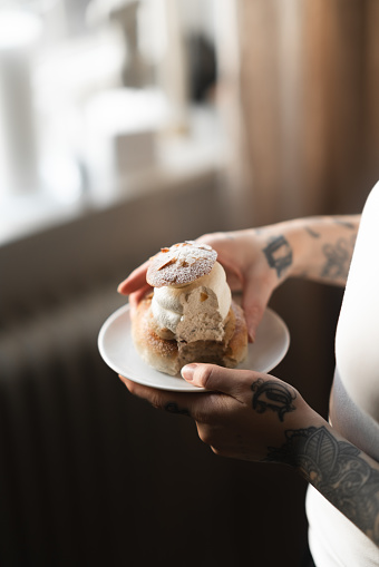 A woman is standing by her window at home, eating a semla pastry. A variation on the traditional Swedish dessert known as Semla, always made and eaten on Fat Tuesday every year. A luxurious looking dessert with almond paste, whipped cream and decorated with chopped almonds and powdered sugar.