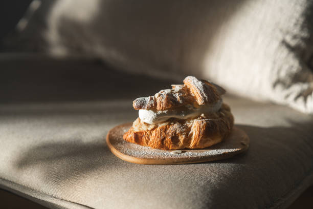 A croissant-semla served on a wooden plate. Placed on the sofa at home. A variation on the traditional Swedish dessert known as Semla, always made and eaten on Fat Tuesday every year. A luxurious looking dessert with almond paste, whipped cream and decorated with chopped almonds and powdered sugar.