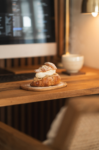 A croissant-semla served on a wooden plate in front of the computer inside the home office. A variation on the traditional Swedish dessert known as Semla, always made and eaten on Fat Tuesday every year. A luxurious looking dessert with almond paste, whipped cream and decorated with chopped almonds and powdered sugar.