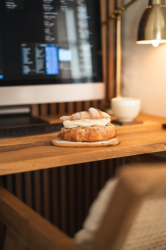 A croissant-semla served on a wooden plate in front of the computer inside the home office. A variation on the traditional Swedish dessert known as Semla, always made and eaten on Fat Tuesday every year. A luxurious looking dessert with almond paste, whipped cream and decorated with chopped almonds and powdered sugar.