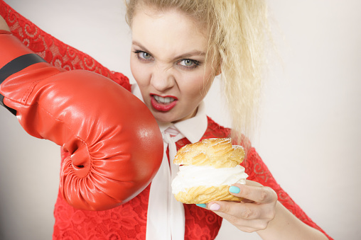 Dieting, weight loss concept. Funny blonde woman holding yummy choux puff cake with whipped cream and boxing glove, fighting off bad food. On grey