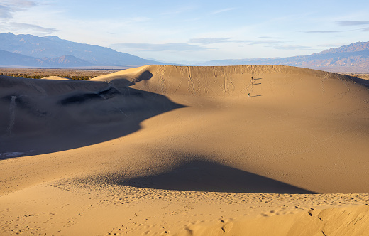 Beneath the tranquil pale blue sky at Mesquite Flat Sand Dunes, the warm hues of the sand create a serene landscape. Unknown tourists, mere specks in the vastness, stroll in the distance, leaving subtle footprints in the desert