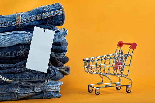 A stack of blue jeans with a white blank tag on a yellow background. Shopping trolley. Sales consept. Heap of stylish trendy denim pants.