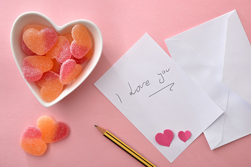 Note written I love you on white paper on a pink background with pencil and white ceramic container with heart-shaped sweets. Top view.