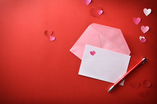 White empty sheet with red pen and open pink envelope on red romantic background with heart cutouts. Top view.