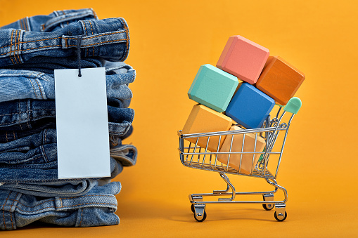 A stack of blue jeans with a white blank tag on a yellow background. Shopping trolley with multi-colored cubes. Sales word written on cubes. Sales consept. Heap of stylish trendy denim pants.