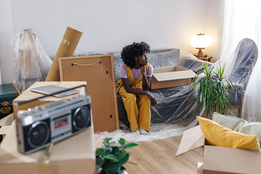 Photo of a young woman sitting on the couch in the living room between half-packed boxes while moving out of her old home.