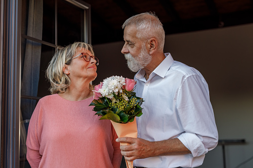 Portrait of romantic mature man giving his loving wife a bouquet of flowers while they are standing at the balcony door.