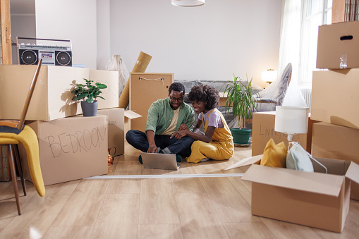 Young family feeling excited to move into a new house. They are using a laptop while sitting on the floor surrounded by cardboard boxes.