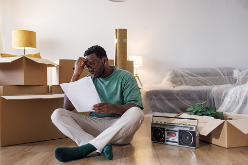 A young African American man with a worried facial expression is sitting on the floor of his new apartment. It looks like he is having a headache while reading a real estate contract.