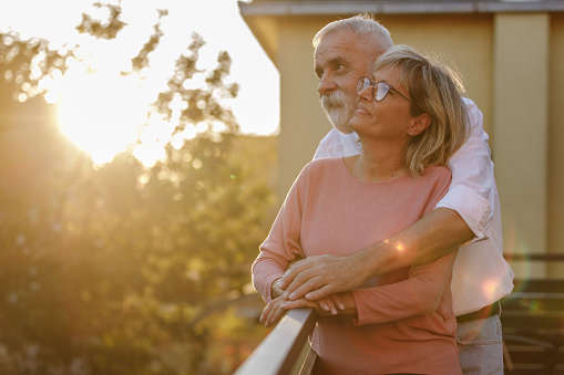 Portrait of happy mature couple standing on the balcony during golden hour, embraced, having a cuddle and bonding moments.
