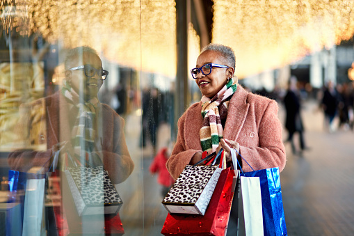 Smiling Black woman in late 50s dressed in warm clothing, holding bags, standing beneath overhead glittering lights and admiring retail merchandise.