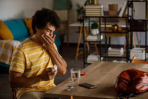 Portrait of mid adult man sitting at his dining table, taking a pill to ease his painful headache.