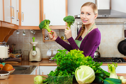 Woman in kitchen with many green leafy vegetables, fresh produce on counter. Young housewife holding broccoli in hand. Healthy eating, cooking, vegetarian food, dieting and people concept.