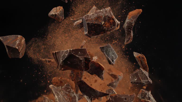 Super slow motion shattered bits and pieces of sweet milk chocolate and cocoa powder tossed in the air on black background