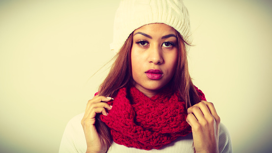 Winter clothing, fashion concept. Beautiful young woman wearing red woolen scarf white cap. Mixed race girl in wintertime clothes portrait, studio shot