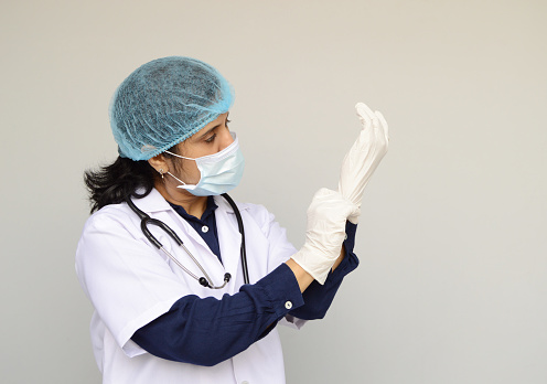 Horizontal photograph of one female doctor with blue shirt, white lab coat, stethoscope getting ready by putting on surgical gloves in her hand over grey background with copy space for text.