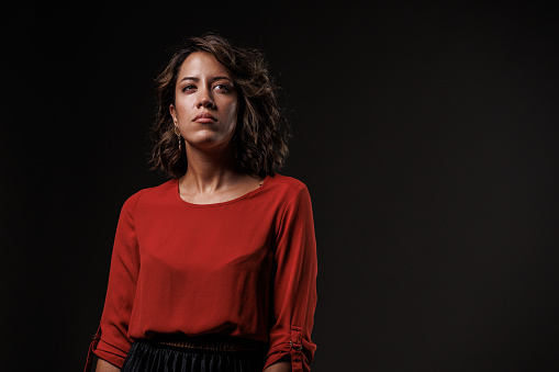 Portrait of unsure young woman, standing against black background, looking away, confused, contemplating.