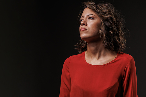 Portrait of unsure young woman, standing against black background, looking away, confused, contemplating.