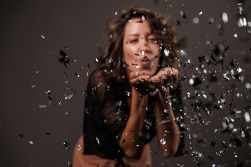 Candid shot of cool young brunette, standing against dark gray background, blowing a handful of silver colored confetti towards camera.