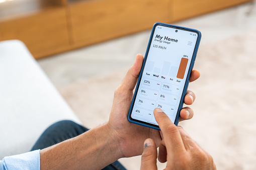 Close-up of hands holding a smartphone to monitor energy usage at his house, tracking day and month data with a focus on energy conservation due to global warming concerns.