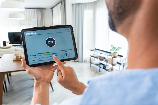 A handsome mid-adult man utilizes voice commands on his tablet's mobile app to turn off lights, adjust brightness, and set a comfortable temperature in his residence, showcasing the seamless integration of home automation.