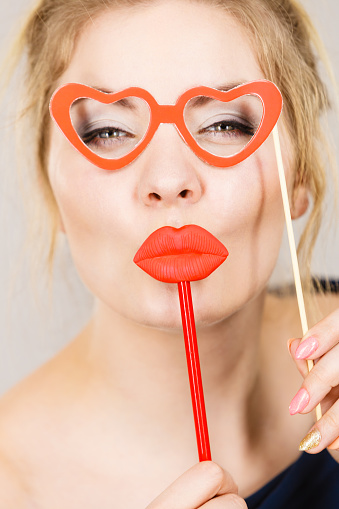 Happy blonde woman holding carnival accessories on stick fake red lips and paper heart shaped glasses, having fun. On grey wall background