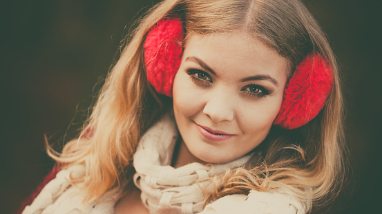 Portrait of pretty smiling fashionable woman in fall forest park. Happy gorgeous young girl in red earmuffs and sweater pullover. Autumn winter fashion.