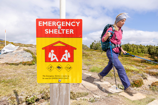 Ensuring safety on the trail, a woman hikes with determination, her path guided by the presence of an emergency shelter sign. A harmonious blend of preparedness and adventure unfolds as she navigates the great outdoors.