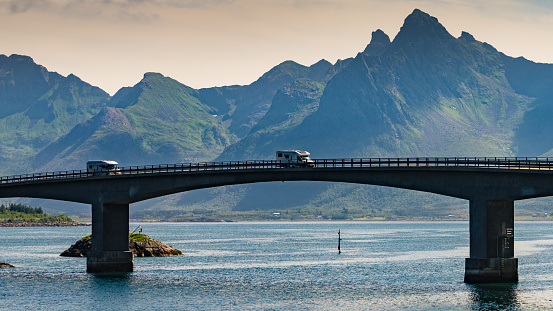 Norwegian scenic landscape on Lofoten archipelago. Camper vehicle on road and bridge connecting islands over sea. Mountain peaks in distance.