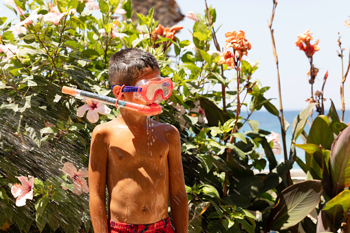 Unrecognizable little boy wearing swimming goggles is sprayed with a hose in garden.