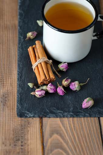 Herbal tea in an enamel mug with ingredients like rose flowers and cinnamon on wooden table.