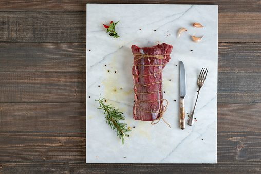 Directly above view of stringed beef on marble background with garlic, rosemary and rustic silver fork and knife.