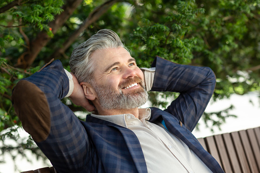 A 45 year old caucasian male in a dark blue blazer and white t-shirt, relaxing outdoors with a contented smile, exuding a serene and happy mood.