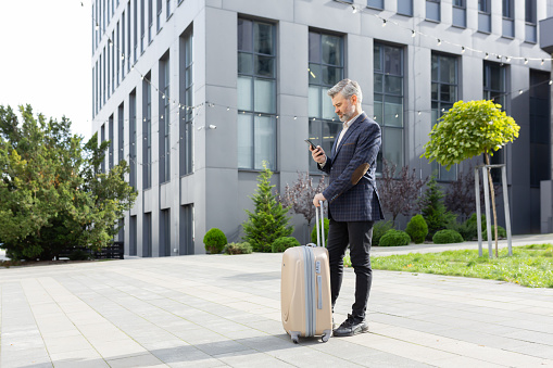 Mature businessman in suit standing with luggage, engrossed in his smartphone outside a contemporary office structure.