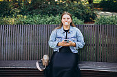 Young hipster female relaxing on the bench in the park.. Model blowing bubble with chewing gum. Happy 30s Woman looking at camera. Summer time