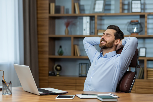 Content mature businessman taking a break, relaxing with hands behind his head in a well-organized home office setting.