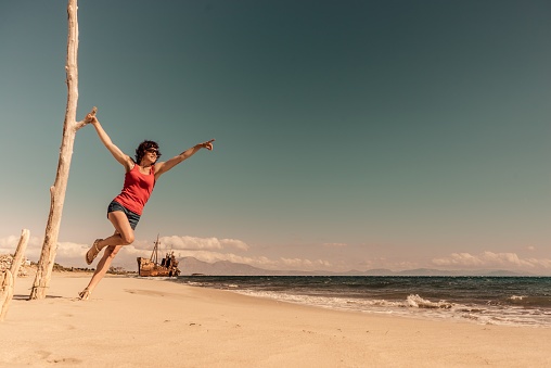 Playful woman having fun while jumping high-up on the beach with her arms raised. Copy space.