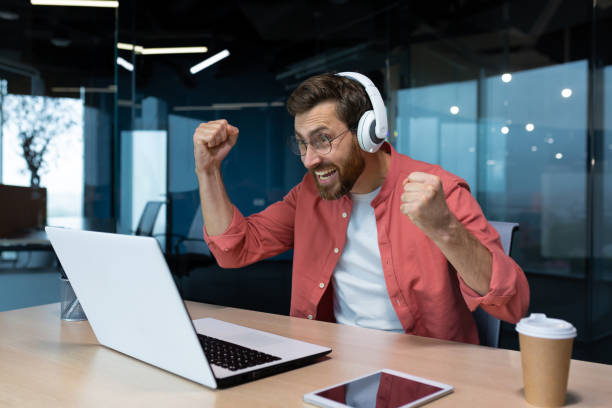 istock Man at the workplace inside the office is cheering watching a sports match on the computer, joyful in headphones, satisfied with the result of the sports team 1960755968
