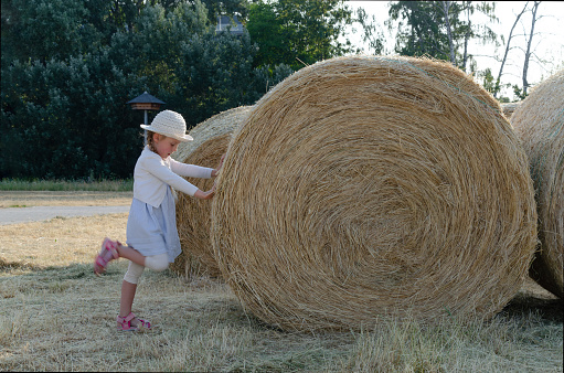 little girl next to big bale of hay. She is wearing a white hat and long pigtails. Hay is rolled up in a bale.