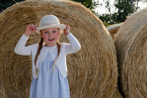 little girl next to big bale of hay. She is wearing a white hat and long pigtails. Hay is rolled up in a bale.