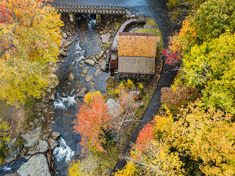 An aerial view of the grist mill at Babcock State Park in West Virginia shows a path decorated in fallen leaves contouring the mill and the gently flowing creek, surrounded by colorful autumnal foliage.