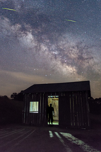 A person stands in silhouette backlit in the entryway of a dark shack inspecting the mysterious night with a clear sky, Milky Way and firefly above.