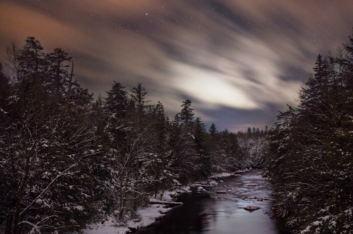 Clouds streak across the West Virginian night sky passing over the Blackwater River and the snow covered pines below in this long exposure.