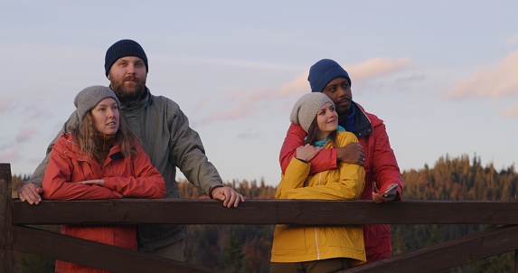 Multiethnic couples stand near wooden fence, look at beautiful sunset enjoying nature during trip. Group of tourists or hiking buddies rest after hike in mountains. Outdoor enthusiasts. Slow motion.