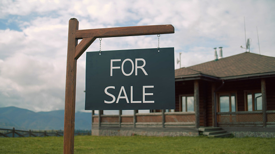For sale sign on beautiful green lawn. Modern house for sale in mountains with amazing landscapes. Stylish property with traditional architecture. Concept of residential property on sale.