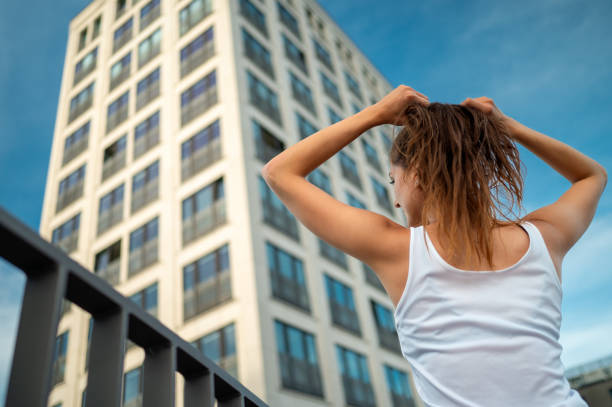 Medium shot of a young caucasian woman against a skycraper binding her hair Low angle perspective from behind of an attractive young woman in a white tank top against a skycraper binding her hair low viewing point stock pictures, royalty-free photos & images