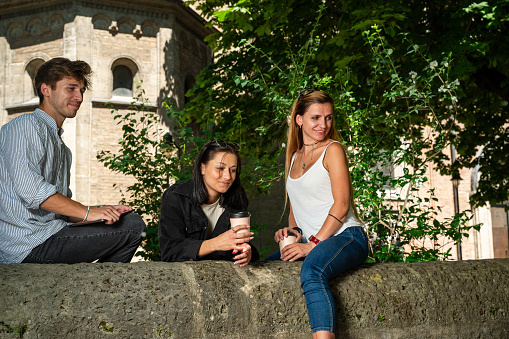 Three young adults sitting and leaning on a small wall and holding coffee-to-go in a city park look at something away from camera with a church on the background on a bright sunny day