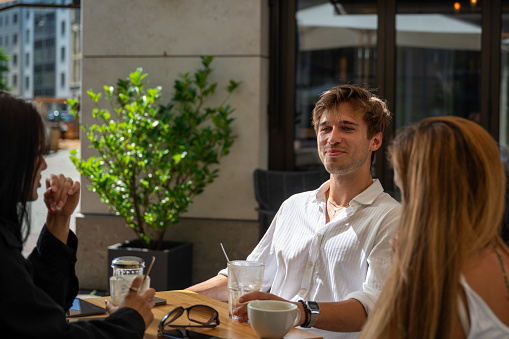 Group of young people wearing sommer garments sit at a bar table with coffee and smiles enjoying the conversation in a bright sunny day
