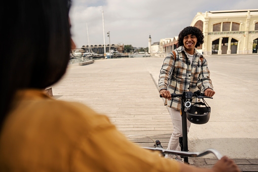 A happy interracial visitor on a rental electric scooter is meeting his friend on a dock during his visit to a coastal Europe city.